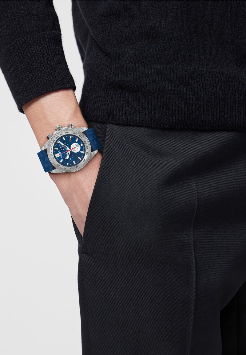 Personne portant une montre-bracelet avec un boîtier en argent, un cadran bleu et un bracelet bleu, main placée de manière décontractée dans la poche d'un pantalon noir, pull noir.