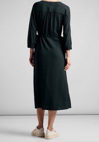 Vestito verde scuro fino al ginocchio con maniche a tre quarti e schiena arricciata. Presenta una cintura in vita e un tessuto testurizzato. Indossato con sneakers bianche.