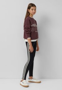 Bordowy sweatshirt z białą grafiką NYC, nałożony na biały T-shirt. Czarne legginsy z białymi paskami po bokach. Białe sneakersy z brązowymi podeszwami.