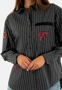 Donna che indossa una camicia nera a righe con bottoni, con una "F" rossa ricamata sulla tasca e una toppa a forma di rosa sul maniche sinistro, con le mani giunte.