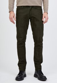 Oliivinvihreät mokkataskut, joissa on kapea leikkaus, vetoketjukiinnitys, viisi taskua ja sileä pinta, yhdistettynä mustiin nauhakengiin.