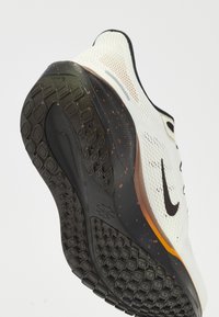 White athletic shoe with a mesh upper, featuring a black swoosh logo and multicolored specks. The sole has a textured black design for grip.