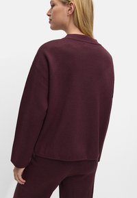 Pulover tricotat de culoare burgundă, cu croială lejeră, umeri lăsați și guler rotund, având o textură netedă și cusături minime.