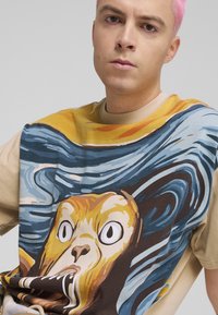 Kortærmet t-shirt med et stort grafik af et bjørnes ansigt på en blå og gul abstrakt baggrund, med en beige ærme. Materialet er en bomuldsblanding.