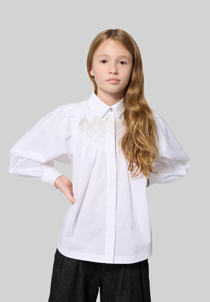 White long-sleeve shirt with a collared neck, featuring a zigzag embroidery detail on the chest and puffed sleeves, paired with black trousers.