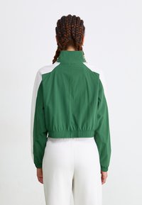 Veste verte courte avec des panneaux d'épaule blancs, une taille élastique, un col haut et des manches longues, fabriquée en matériau léger.