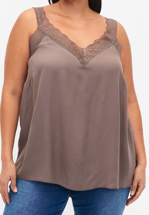 Frau trägt ein ärmelloses taupefarbenes Oberteil mit Spitzenbesatz am Kragen und blue Jeans, steht vor einem schlichten Hintergrund.