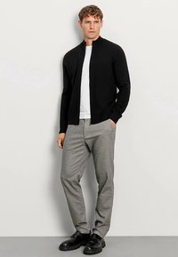 Schwarzer Reißverschluss-Pullover, weißes T-Shirt, graue Hose und schwarze Schuhe. Der Pullover hat einen Stehkragen und ein tailliertes Design.