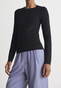Pull en tricot noir avec motif texturé, encolure ronde et poignets ajustés. Associé à un pantalon large lavande clair.