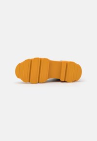 Semelle de chaussure avec un épais caoutchouc jaune, présentant de larges rainures horizontales et des détails de grip texturé sur un fond blanc.
