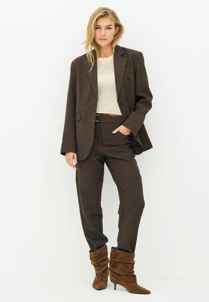 Femme portant un tailleur marron foncé avec un blazer ample et un pantalon large, un crop top en maille crème, et des bottines à talons en daim marron décontractées, debout devant un fond blanc.