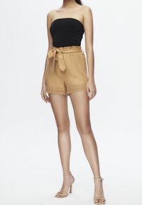 Top nero senza spalline abbinato a pantaloni shorts alti color beige con vita arricciata e rifiniture in pizzo. La modella indossa sandali con tacco trasparente.
