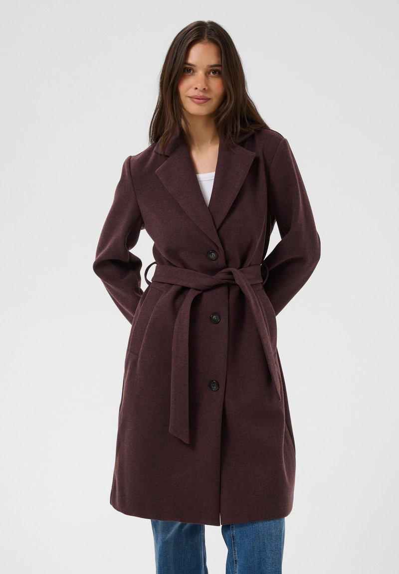 Femme portant un manteau ceinturé violet foncé avec des boutons sur un jean bleu, debout les mains dans les poches, regardant devant avec une expression neutre.