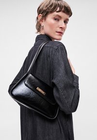 Μαύρη δερμάτινη τσάντα ώμου με ορθογώνιο σχήμα, γυαλιστερή υφή και χρυσές λεπτομέρειες. Διαθέτει μπροστινό καπάκι και μακρύ ρυθμιζόμενο λουρί.