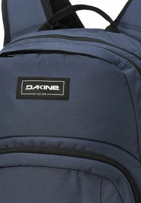 Dakine-ryggsäck i mörkblått tyg har en slät yta, logopatch och svarta dragkedjor, med vadderade axelremmar upptill.