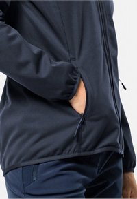 Veste softshell marine avec une poche latérale zippée ; tissu lisse ; poignets élastiques ; design ajusté ; zips et détails de couture visibles.