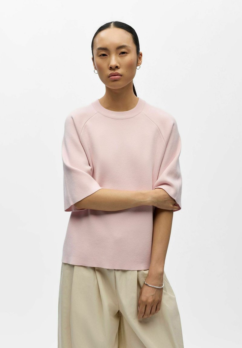 Femme aux cheveux bruns attachés portant un pull léger rose à manches courtes et un pantalon beige, debout avec un bras croisé sur l'autre.