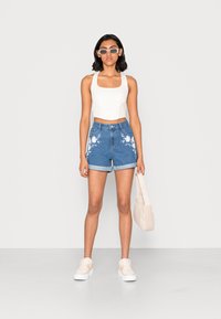 Λευκό κοντό τοπ, τζιν σορτς με λευκή λουλουδάτη κεντήματα, διπλωμένα μανίκια και λευκά αθλητικά παπούτσια; κρατάει μια μπεζ τσάντα υφής.