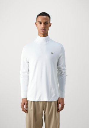 Chemise à col roulé blanche à manches longues en matériau doux, avec un logo vert Lacoste sur la poitrine, associée à un pantalon beige clair.