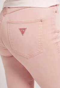 Różowe dżinsy z denimu z trójkątnym wzorem kieszeni i czerwonym logo "GUESS". Materiał ma gładką teksturę i dopasowany krój.