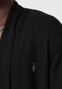 Mustat, ribbineuletakki avointa etuosaa, jossa on hienovarainen vuohen kallo -symboli vasemmalla puolella. Teksturoitu kangas, rento istuvuus.