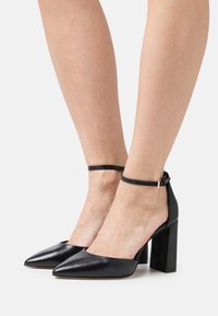 Chaussures à talons hauts en cuir noir avec un bout pointu, une bride de cheville et un talon large, présentant une texture lisse et un design minimaliste.