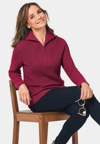 Bordeaux trui met een half-rits kraag, geribbelde textuur en lange mouwen, in combinatie met donkere jeans en ronde zonnebrillen, zittend op een houten stoel.