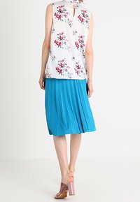 Blouse sans manches à motifs floraux en blanc avec des détails roses et verts, associée à une jupe plissée turquoise et des mules à talons transparents.