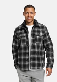 Sort og hvid ternet flanneljakke med to brystlommer, knaplukning og en blød tekstur, båret over en hvid T-shirt med rund hals.