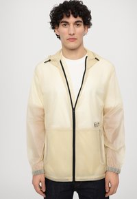Light yellow, semi-transparent jacket with a front zipper, elastic cuffs, and drawstring hood. Features black branding on the chest.