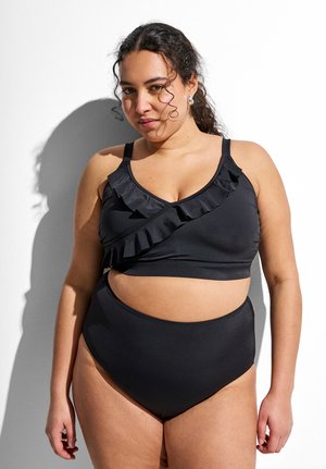 Kučeravá žena stojí pri bielej stene, nosí čierny volánikový krátky plavkový top a bikiny s vysokým pásom.