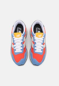 Punaiset ja siniset mokka- ja verkkanilkkurit, joissa valkoiset nauhat, vaaleanruskeat yksityiskohdat, mustat detaljit ja keltainen logo-tagi kielellä.