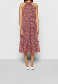 Halterneckklänning i rött blommönster med flerstegs fåll, tillverkad av lätt tyg. Stylad med vita skor.