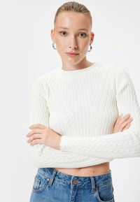 Maglione bianco a coste con motivo a trecce, scollo rotondo e maniche lunghe. Abbinato a jeans in denim blu a vita alta.
