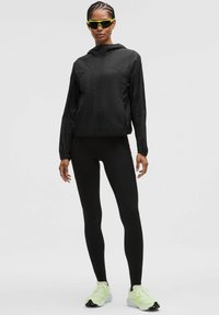 Sort letvægtsjakke med lynlås og hætte, parret med tætsiddende sorte leggings og klare grønne sportssko. Solbrillerne har en neongrøn detalje.