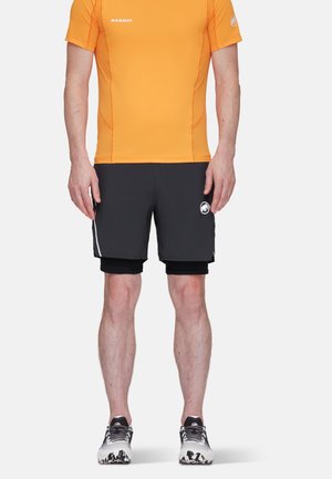 Man draagt een feloranje atletisch shirt met korte mouwen, zwarte dubbele laag shorts en hardloopschoenen, staand tegen een effen witte achtergrond.