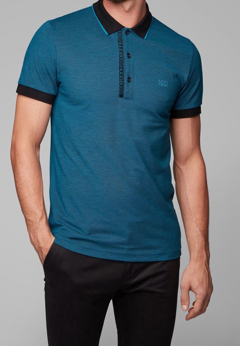 Homme portant un polo sarcelle avec col et poignets noirs, boutons noirs et pantalon noir, debout devant un fond gris.