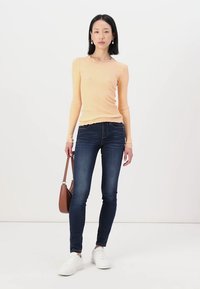 Lange mouwen geribbelde top in licht perzik, gecombineerd met strakke donkerblauwe jeans en witte sneakers. Bruine schoudertas maakt de look compleet.