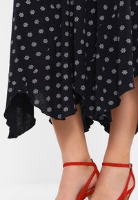 Black dress with a floral white pattern, featuring a high-low hemline and lightweight fabric. Red strappy sandals accentuate the look.