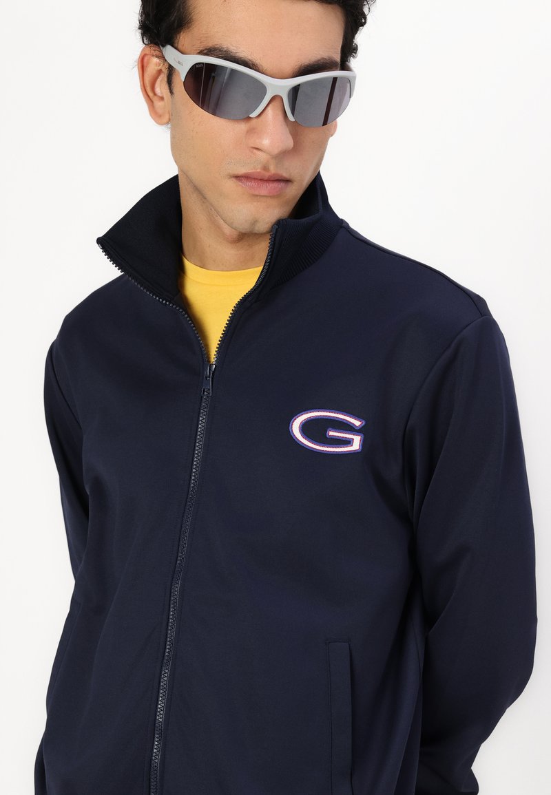 Chaqueta navy con cremallera y cuello alto, logo bordado; el modelo lleva gafas de sol grises y una camiseta amarilla debajo, mostrando un aspecto deportivo.