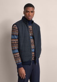 Dunkelgraue Weste mit hohem Kragen, gestepptem Stoff und Reißverschluss. Darunter ein gemusterter Pullover in Brauntönen, Blau und Weiß.