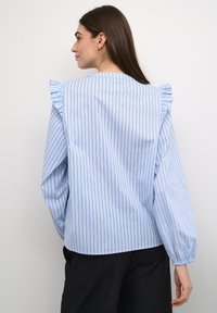 Camicia azzurra con strisce verticali bianche e marroni, spalle arricciate, maniche lunghe e polsini elastici. Tessuto di cotone liscio.