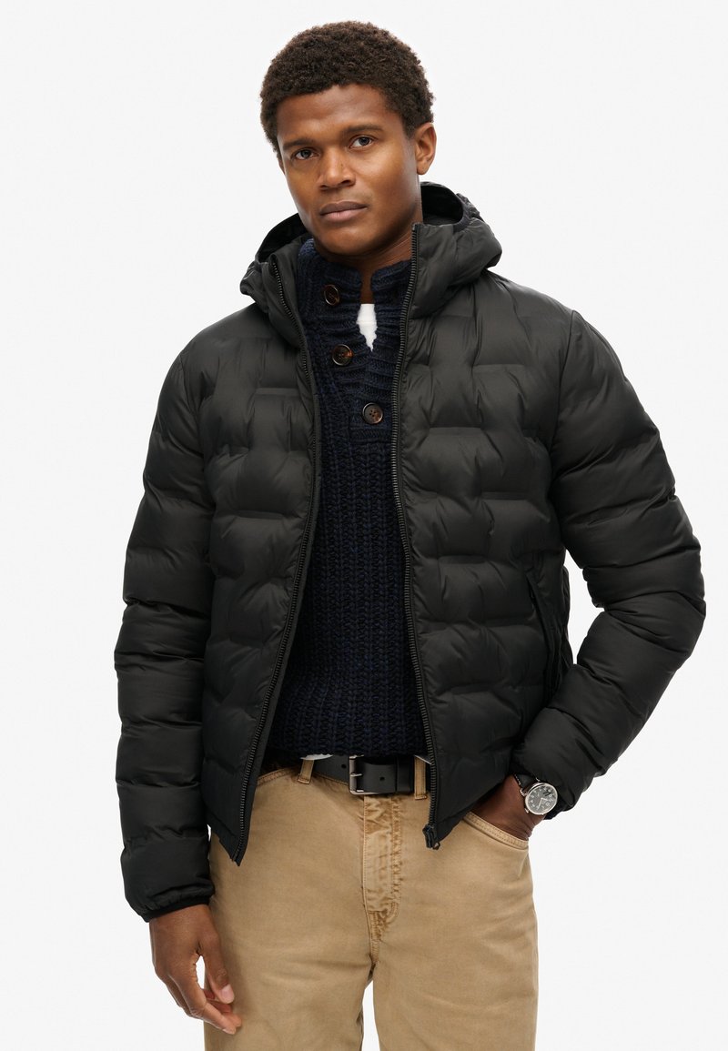 Chaqueta de plumas negra con capucha, diseño acolchado, cremallera frontal, llevada sobre un suéter de punto azul oscuro. Combinada con pantalones beige y un reloj.