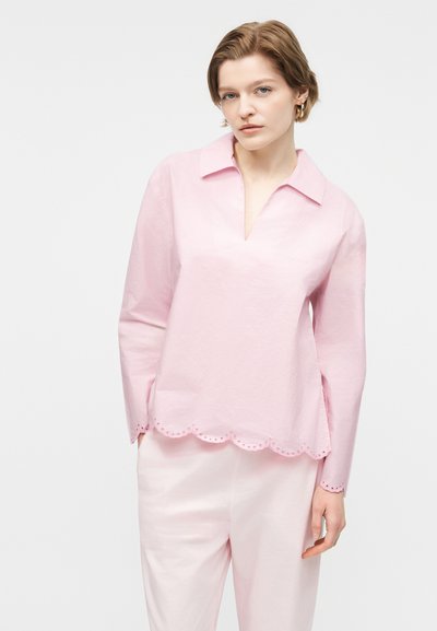 Fiatal nő világos rózsaszín, hullámos szegélyű hosszú ujjú blúzban és fehér nadrágban, sima fehér háttér előtt áll.