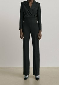 Svart skräddarsydd jumpsuit med långa ärmar, sjalkrage, midjeskärp och vida byxor, tillsammans med silvriga klackskor.