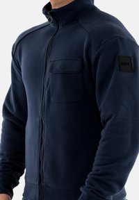 Giacca in pile blu navy con zip, colletto alto, tasca frontale e tessuto strutturato. Presenta una patch del logo sulla manica sinistra.