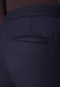 Marineblauwe broek met een gestructureerde stof, een achterzak met een gestikte opening, en een gladde tailleband.