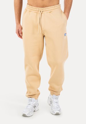 Pantaloni della tuta beige con vita e polsini elastici, piccolo logo NBA sulla coscia sinistra, indossati con scarpe New Balance bianche, mani in tasca.