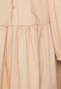 Robe beige à manches longues avec taille froncée, devant boutonné et poignets élastiques, confectionnée dans un tissu léger.