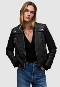 Giacca biker in pelle nera con dettagli trapuntati sulle spalle, hardware silver, tasche con zip e indossata sopra un maglione nero con bottoni.